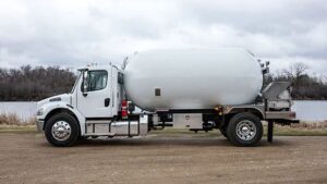 A Blueline QX Bobtail propane delivery truck, shown from a side view. The truck is a large industrial vehicle with a prominent cylindrical propane tank mounted on its chassis. A hose reel and control panel are visible at the rear of the tank. The tank and truck cab appear to be a dark color.